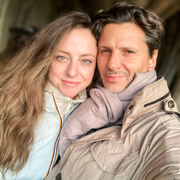 The image depicts a man and woman posing for a selfie, with the woman on the left and the man on the right, both wearing jackets and standing in front of a blurred background.