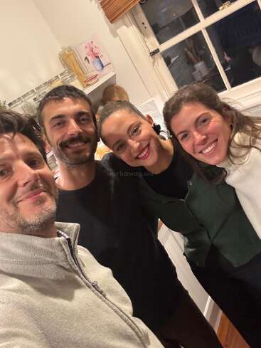 Four people are smiling together for a selfie in a cozy kitchen. They look happy and relaxed, enjoying each other's company in a warm atmosphere.