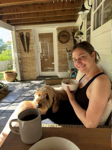 Une femme sourit, assise sous un porche, en mangeant dans un bol. Un chien en peluche se repose sur ses genoux. Un café et la lumière du soleil complètent ce décor chaleureux.