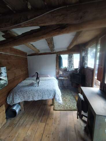 Una acogedora habitación rústica con vigas y suelos de madera, una cama con ropa de cama floral, un gato de pie sobre la cama, un escritorio y una ventana.