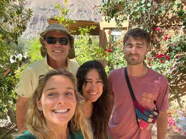 Quatre personnes sourient pour une photo en plein air dans un jardin luxuriant, ensoleillé, avec des fleurs et de la verdure. Des montagnes sont visibles en arrière-plan, créant une atmosphère joyeuse.
