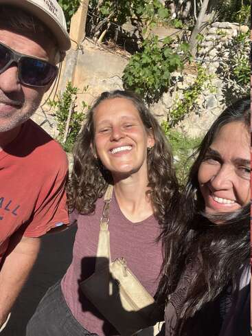 Trois personnes sourient au soleil. Elles semblent heureuses et proches, profitant d'un cadre extérieur avec de la verdure et des murs en pierre en arrière-plan.