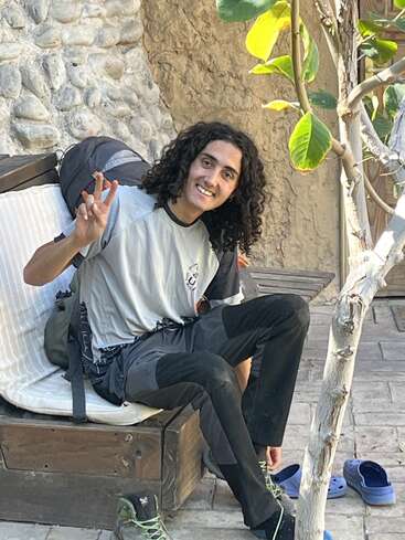 Un jeune aux cheveux bouclés est assis sur un banc en plein air, souriant et faisant un signe de paix. Ils portent des vêtements de randonnée, avec des chaussures à proximité et de la verdure autour.