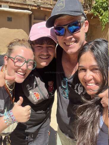 Quatre personnes sourient au soleil, posant côte à côte. Elles portent des vêtements décontractés et des lunettes de soleil, et rayonnent de bonheur. Une personne lève le pouce. Cadre extérieur.
