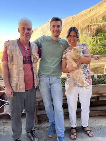 Trois personnes se tiennent côte à côte, souriant à l'extérieur, avec des montagnes en arrière-plan. La femme à droite tient dans ses bras un chat couleur crème, qui profite de l'instant présent.