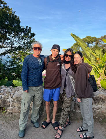 Quatre personnes se tiennent côte à côte en plein air, souriantes et habillées de manière décontractée. Ils sont entourés d'une végétation luxuriante et d'un ciel bleu clair, ce qui crée une atmosphère détendue et joyeuse par une journée ensoleillée.