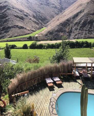 A relaxing pool area with lounge chairs overlooks lush green fields and vineyards, surrounded by dry, rugged mountains under bright sunlight, creating a serene atmosphere.