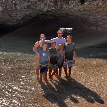 L'image représente une famille de six personnes sur une plage, avec une grande grotte à l'arrière-plan et une étendue d'eau derrière eux.