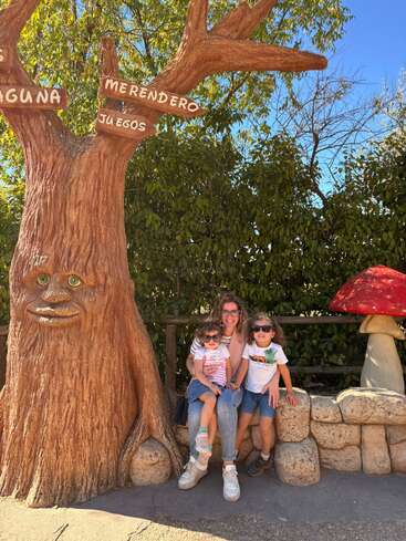 Eine Frau und zwei Kinder sitzen neben einer skurrilen Baumskulptur mit einem Gesicht, bunten Schildern und einem roten Pilz, umgeben von üppigem Grün unter sonnigem Himmel.