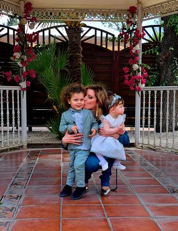 Eine Frau sitzt auf einem Stuhl, hält ein kleines Mädchen und einen kleinen Jungen und posiert für ein Foto in einem Garten mit gefliestem Boden und weißem Geländer.