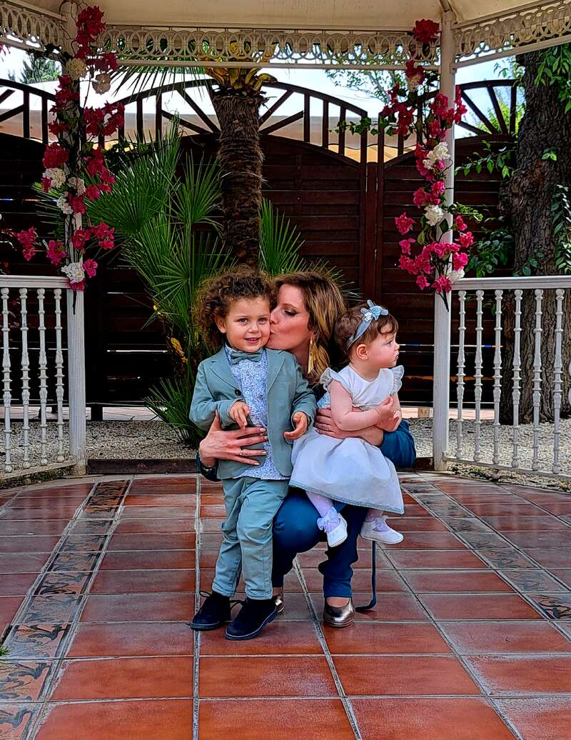 Eine Frau sitzt auf einem Stuhl, hält ein kleines Mädchen und einen kleinen Jungen und posiert für ein Foto in einem Garten mit gefliestem Boden und weißem Geländer.