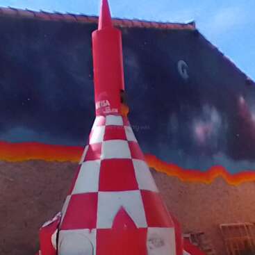 L'image représente une sculpture de fusée à carreaux rouges et blancs contre une peinture murale vibrante, sur fond de ciel bleu avec quelques nuages ondulants.