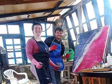 Duas pessoas usando aventais e luvas sorriem enquanto pintam em um estúdio artístico banhado pela luz do sol. Materiais de arte, uma tela colorida e energia criativa preenchem a sala.