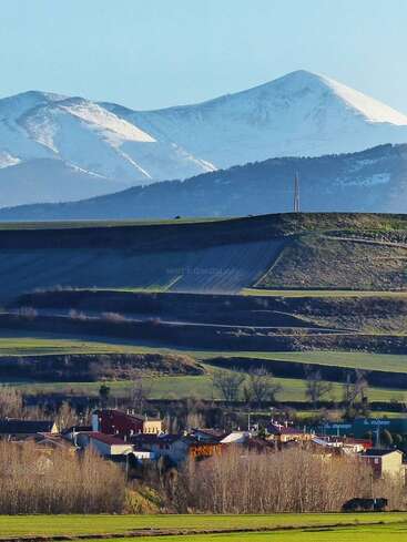 A imagem retrata uma paisagem serena com colinas onduladas, montanhas cobertas de neve e uma pequena aldeia em primeiro plano, banhada pela luz do sol.