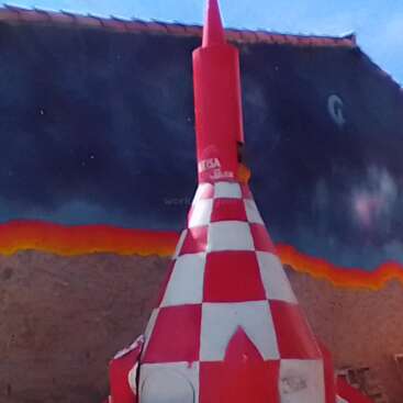 L'image représente une sculpture de fusée à carreaux rouges et blancs contre une peinture murale vibrante, sur fond de ciel bleu avec quelques nuages ondulants.