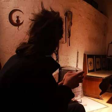 L'image représente une femme assise par terre devant un mur, tenant une tasse, avec une silhouette de chat sur le mur et une chaise en bois à proximité.