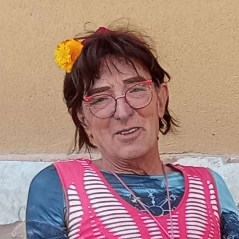 Uma pessoa com cabelo castanho curto, uma flor amarela no cabelo, óculos de armação vermelha e uma blusa colorida está sentada encostada a uma parede bege, sorrindo calorosamente.