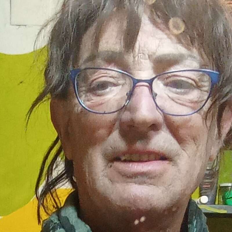 Eine Nahaufnahme einer älteren Person mit kurzen braunen Haaren, einer blauen Brille und einem grünen Schal, die drinnen vor einem gelb-weißen Hintergrund steht.