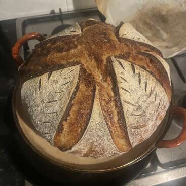 A beautifully baked round loaf of artisan bread sits in a pot, featuring decorative scoring with a rustic, golden crust, dusted lightly with flour.