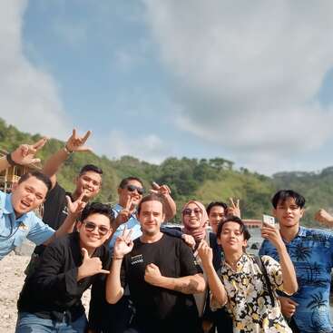 The image depicts a group of 10 people posing for a photo on a rocky beach, with a body of water and a mountainous landscape in the background.