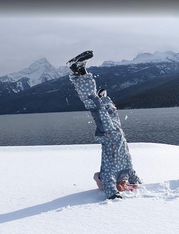 Eine Person in einem blauen Schneeanzug mit weißen Blumen macht einen Kopfstand im tiefen Schnee, mit verschneiten Bergen und einem See im Hintergrund. Verspielte Winterszene.