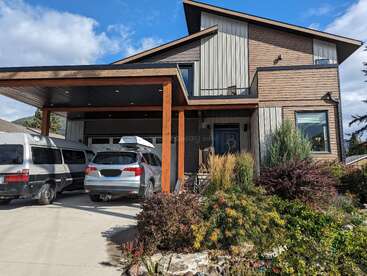 Ein modernes zweistöckiges Haus mit schrägem Dach, Holzfassade und großen Fenstern. Zwei Fahrzeuge sind unter einem Carport geparkt; vor dem Haus gibt es einen üppigen Garten.