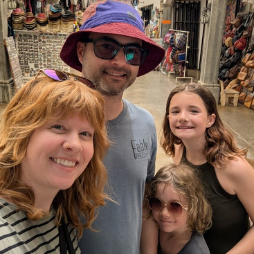 Eine glückliche vierköpfige Familie posiert für ein Selfie auf einem Markt im Freien, umgeben von Hüten, Taschen und Souvenirs, alle lächeln und genießen ihren Tag.