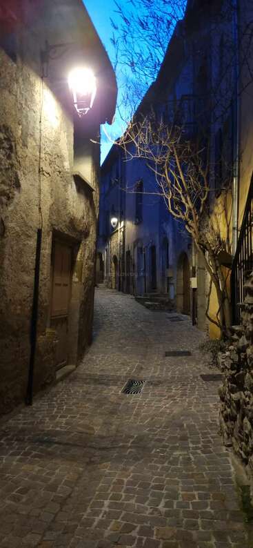 Eine schmale Gasse aus Kopfsteinpflaster schlängelt sich zwischen alten Steinhäusern hindurch, die von Straßenlaternen schwach beleuchtet und von kahlen Bäumen vor einem dämmrigen blauen Himmel eingerahmt werden.