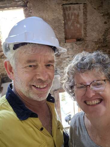 Ein lächelnder Mann in einem gelben Arbeitshemd und einem weißen Schutzhelm steht neben einer fröhlichen Frau mit Brille in einem rustikalen Steingebäude, das gerade gebaut wird.