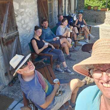 Un groupe de sept personnes se détend en plein air, assis sur des chaises, souriant et prenant un verre ensemble. C'est un endroit ensoleillé et décontracté, avec un bâtiment rustique en pierre à l'arrière-plan.
