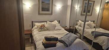 A cozy bedroom with white bedding, two pillows, folded clothes on the bed, wall art, bedside tables, mirrored closets, soft lighting, and tiled flooring.