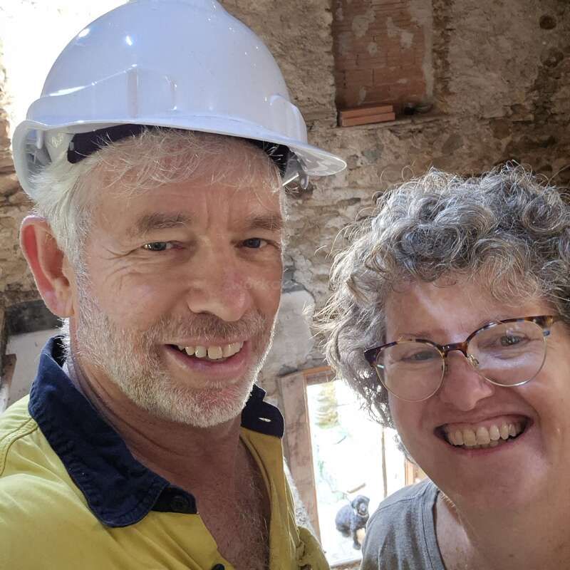 A smiling man in a yellow work shirt and white hard hat stands beside a cheerful woman with glasses, inside a rustic stone building under construction.