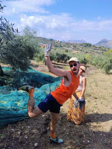 A man playfully poses with one leg up, wearing bright athletic clothing and gloves, while a woman in a hat smiles behind him in a sunny olive grove.