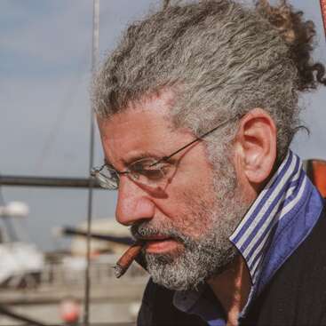 Un hombre de pelo y barba rizados y grises, con gafas, parece pensativo. Tiene un puro en la boca y está al aire libre cerca de un puerto deportivo.