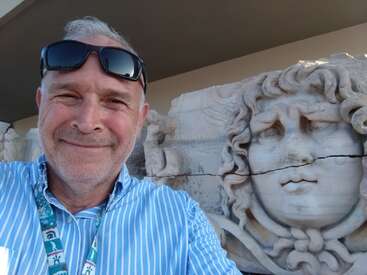 Ein lächelnder Mann in einem blau gestreiften Hemd und mit Sonnenbrille macht ein Selfie neben einer alten Steinskulptur mit einem zerklüfteten, ausdrucksstarken Gesicht und lockigem Haar.