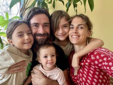 The image depicts a family of five posing for a photo, with the father holding a baby and the mother embracing her two children, all smiling and surrounded by plants.