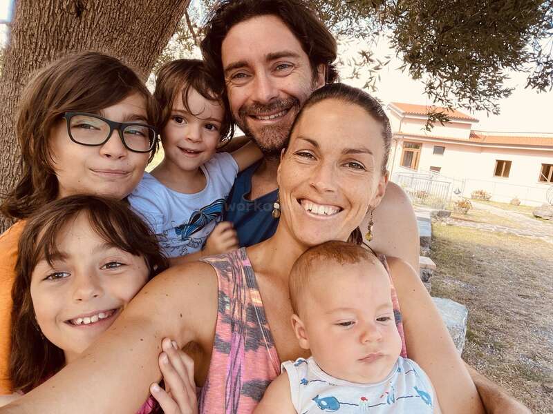 A happy family of six smiles together outdoors under a tree. Parents and four children, including a baby, enjoy a sunny day near a building.