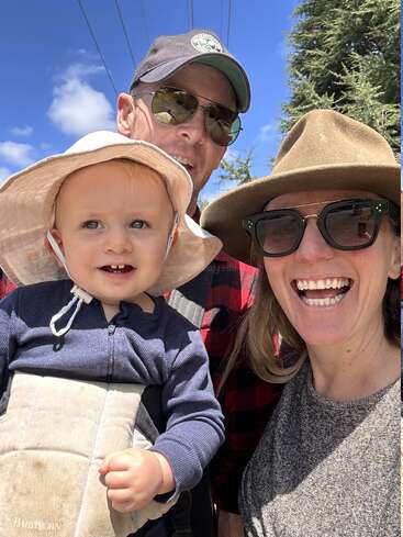 Un selfie de famille joyeux pris en extérieur, montrant deux adultes portant un chapeau et des lunettes de soleil, ainsi qu'un enfant souriant coiffé d'un chapeau de soleil et porté dans un porte-bébé, sous un ciel d'un bleu éclatant.