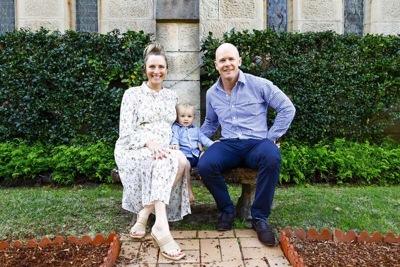 Une famille de trois personnes, tout sourire — une mère, un père et un jeune enfant —, est assise sur un banc en pierre dans un jardin, avec une végétation luxuriante et un bâtiment en pierre en arrière-plan.