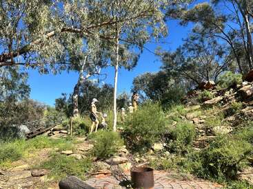 Cuatro niños y un adulto suben por una ladera rocosa y cubierta de hierba, rodeada de árboles, bajo un cielo azul brillante en un día soleado, mientras exploran juntos la naturaleza al aire libre.