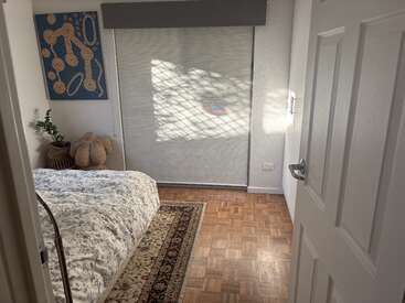 Sunlight filters through a window onto the floor of a cozy bedroom with a patterned bed, woven rug, house plant, abstract artwork, and teddy bear.