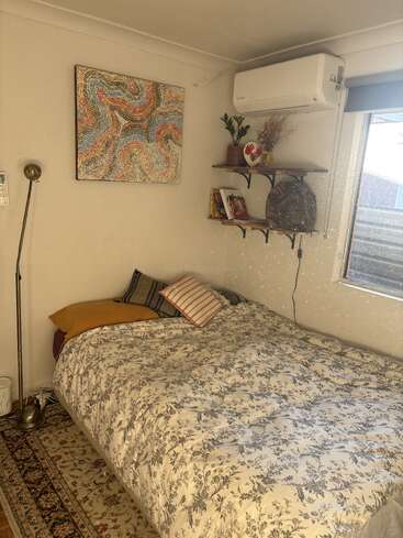 A cozy bedroom with a floral bedspread, patterned pillows, wall art, a standing lamp, shelves with books and decor, air conditioner, and natural light from a window.