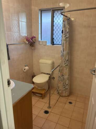 This bathroom features peach tiles, toilet, safety rail, small wooden stool, floral shower curtain, window, towel, toilet paper holder, artificial flowers, and a sink.