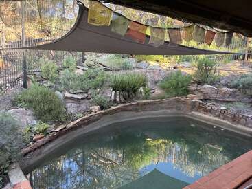 A small, tranquil pool with weathered rock edging sits beneath a shade sail and colorful prayer flags, surrounded by lush greenery and natural stone landscaping.