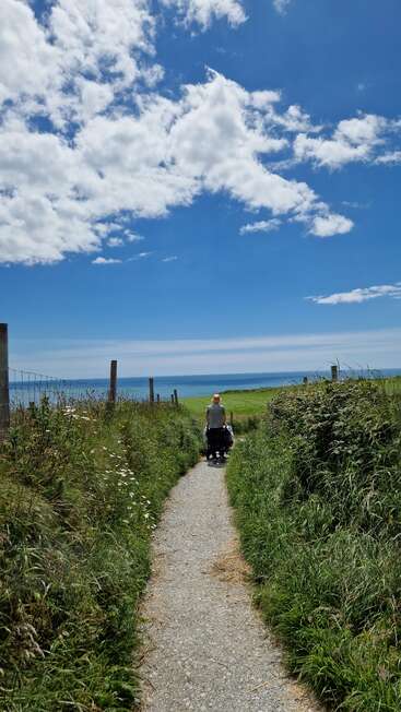 Une personne poussant une poussette marche sur un chemin étroit à travers des herbes hautes en direction de la mer, sous un ciel d'un bleu éclatant parsemé de nuages cotonneux.