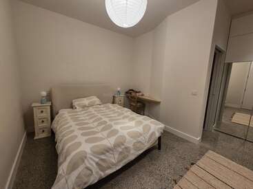 Cette image montre une chambre à coucher minimaliste avec un lit double, une couette à motifs, des tables de chevet, un bureau, une chaise, un placard à miroirs, des murs neutres et un plafonnier rond.