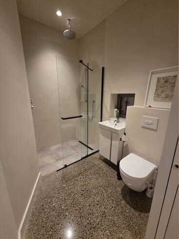 Cette image montre une salle de bains moderne avec une douche à l'italienne, une cloison en verre, des toilettes murales, un petit lavabo, un sol en béton poli et un décor minimaliste dans des tons neutres.