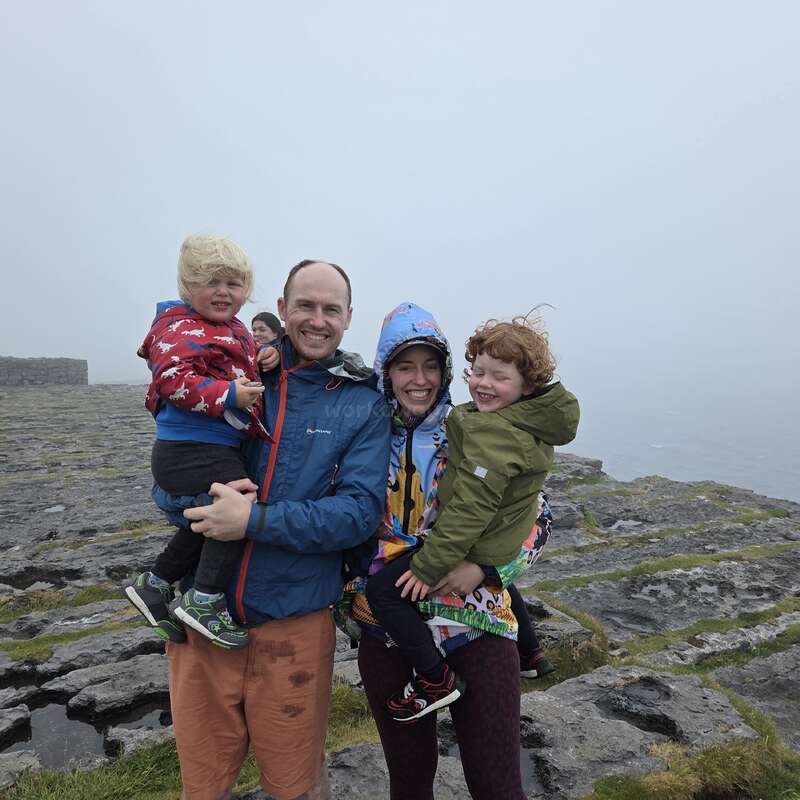 Une famille de quatre personnes, emmitouflées dans des vestes colorées, sourit sur une falaise rocheuse par temps brumeux. La mer et le ciel gris forment l'arrière-plan.