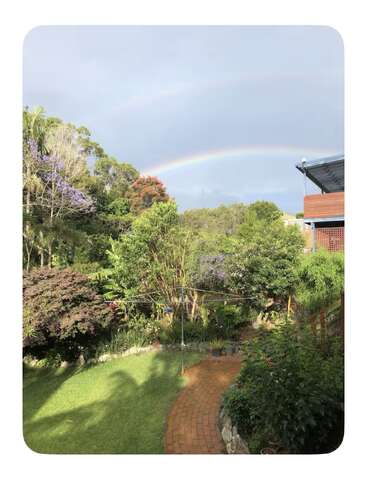 Um belo jardim com vegetação exuberante, flores coloridas e um caminho sinuoso de tijolos. Acima, um arco-íris vibrante se estende por um céu nublado, criando uma cena pacífica.