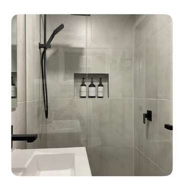 Esta imagem mostra um chuveiro moderno e minimalista com grandes azulejos cinza claro, luminárias pretas foscas, uma prateleira embutida e três garrafas dispostas ordenadamente dentro da prateleira.